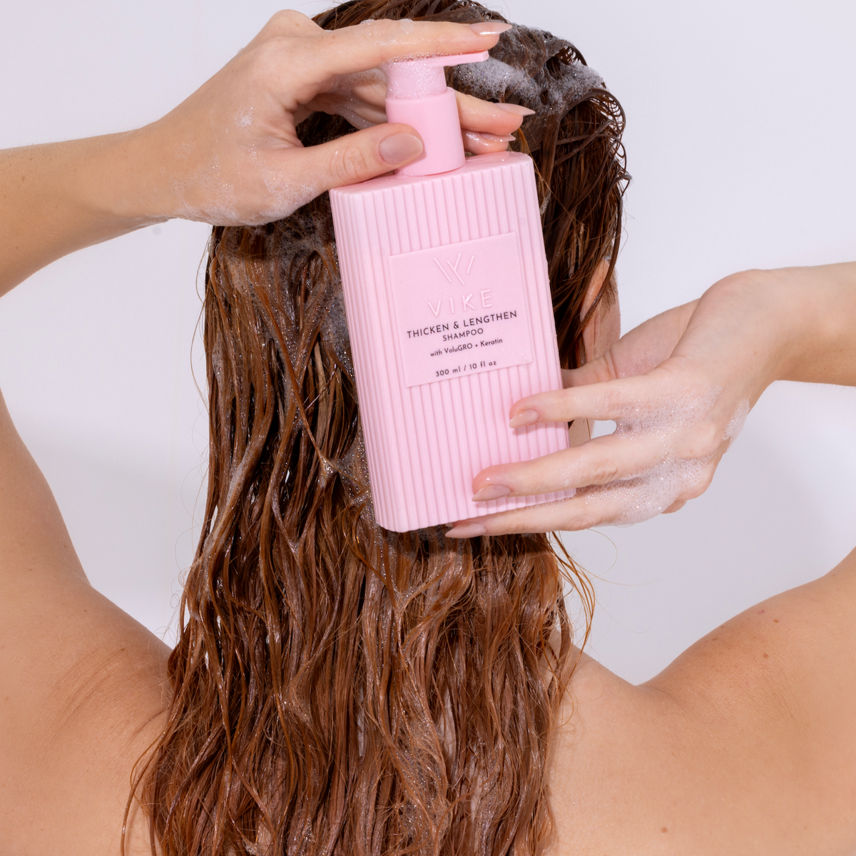 Person applying pink shampoo bottle to wet hair against a white background

